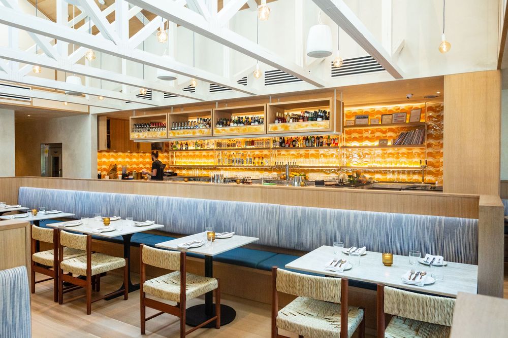 Modern restaurant interior with gray banquette seating and illuminated bar shelving under white industrial ceiling beams.
