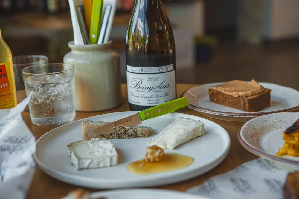 A table setting with cheese plate, wine bottle, and bread accompaniments on white plates with a glass of water.