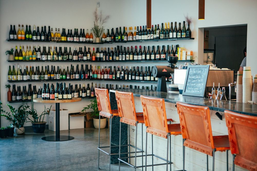 Modern wine bar interior with shelves of bottles and orange leather barstools at a sleek counter area.