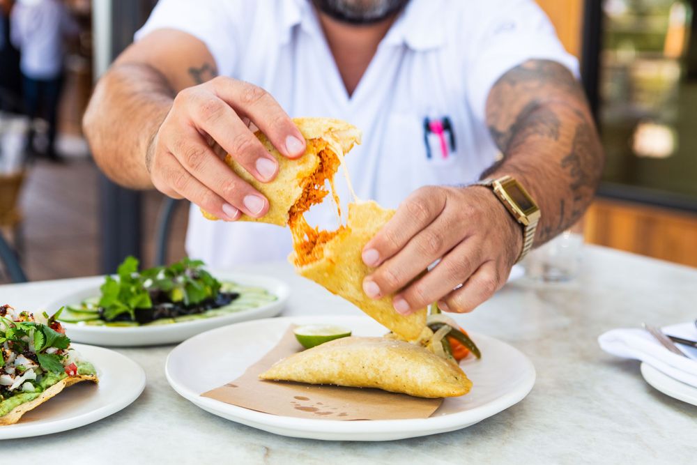 Hands drizzle mustard sauce onto a sandwich at a restaurant table with side dishes visible.