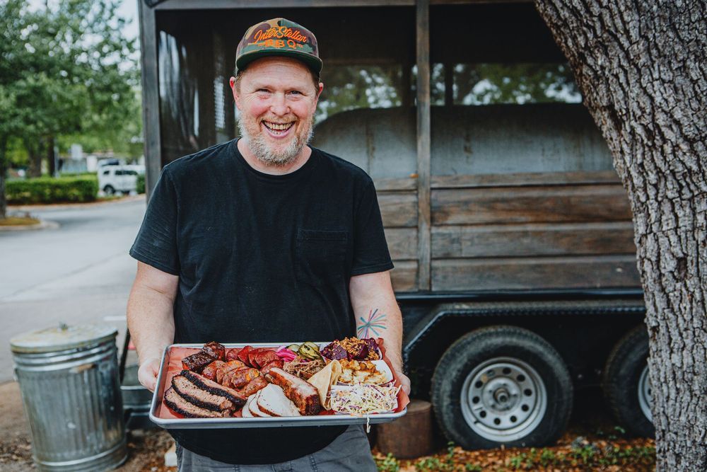 Someone in a black t-shirt and cap holds a tray of smoked barbecue meats and sides next to a smoker trailer.