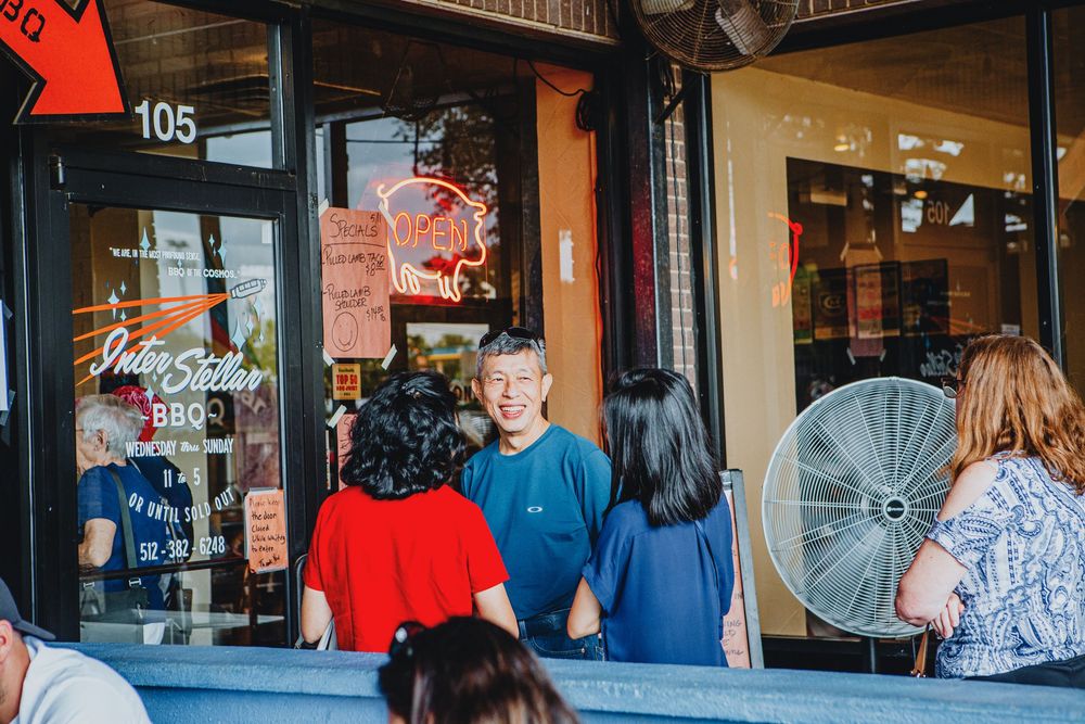 People gather outside a storefront window on a busy street with a red neon sign glowing in the background.