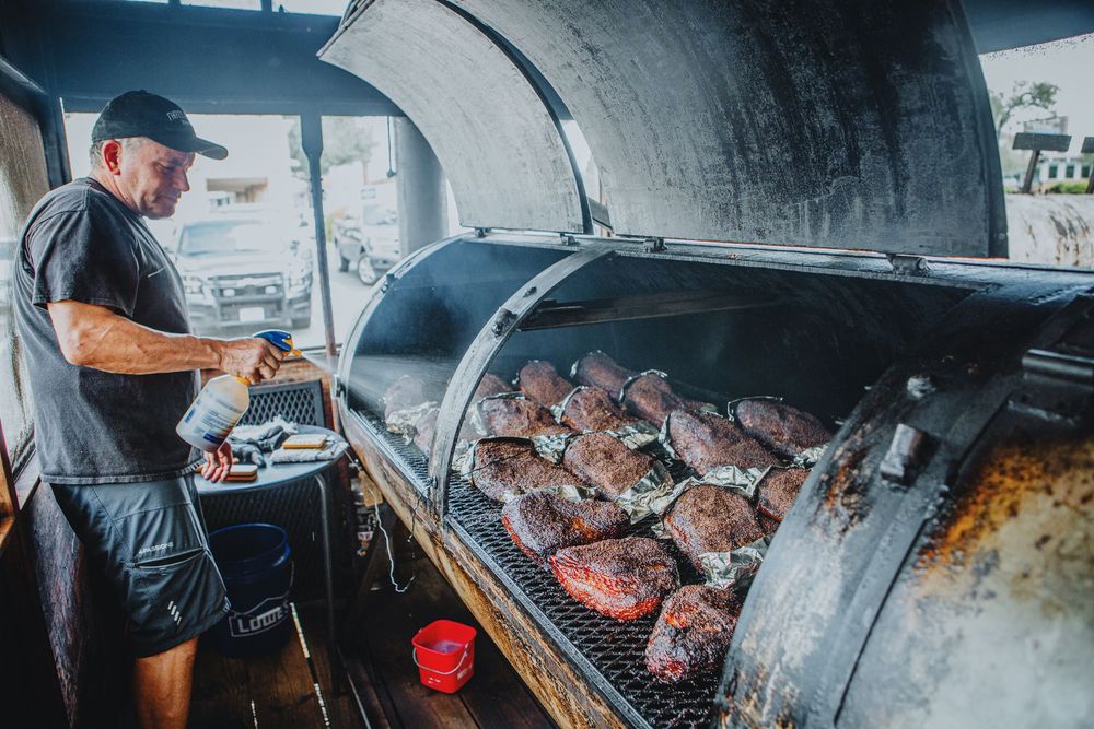 A pitmaster tends to multiple cuts of meat smoking in a large cylindrical barbecue smoker.