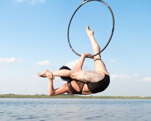 An aerial performer in a black outfit gracefully poses with a lyra hoop over calm water against a blue sky.