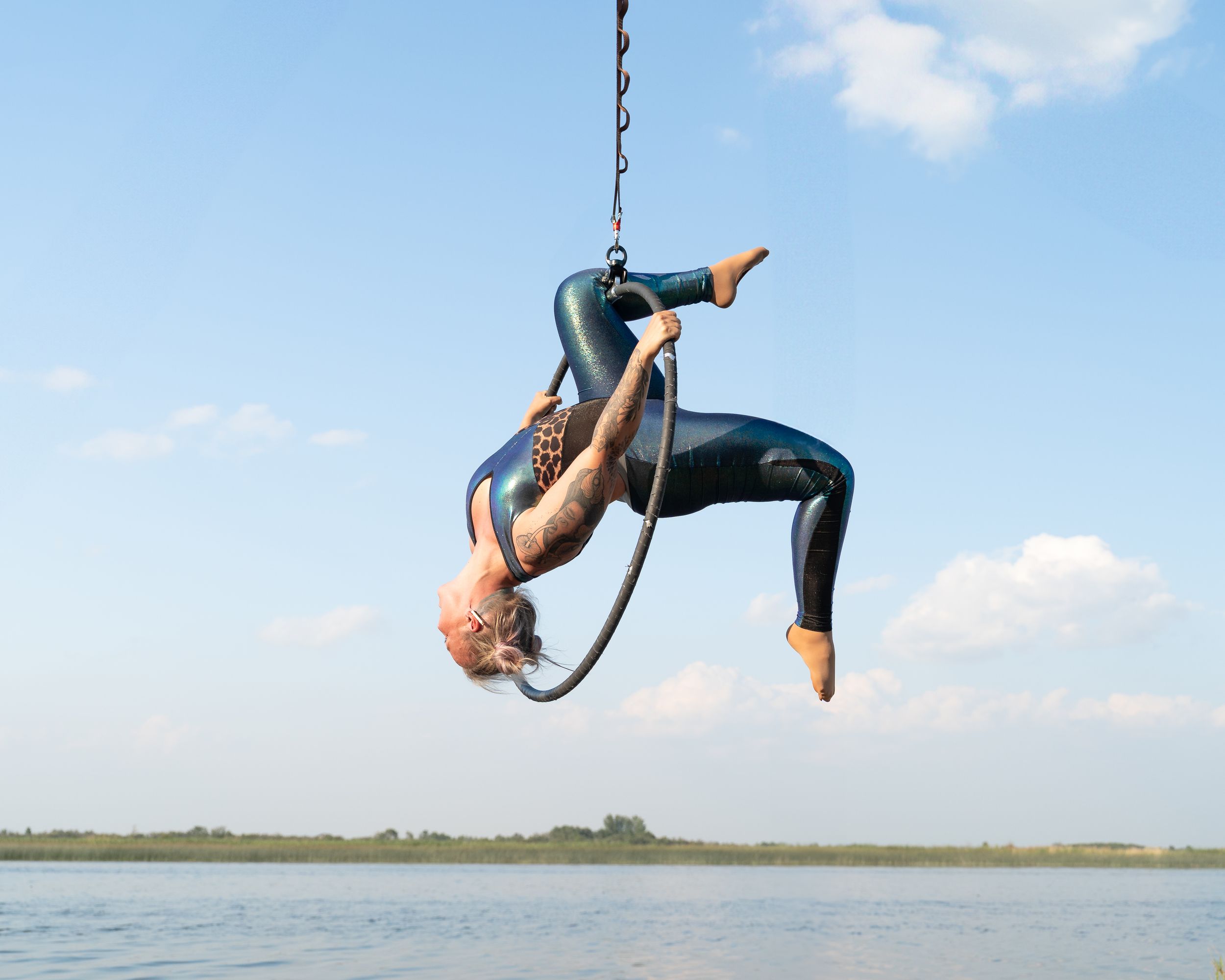 Aerial performer gracefully suspended from aerial hoop over tranquil lake on sunny day in dynamic backbend pose.