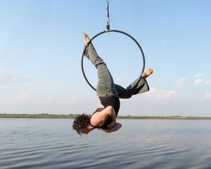 An aerial performer hangs upside down from a suspended hoop over calm water against a blue sky.
