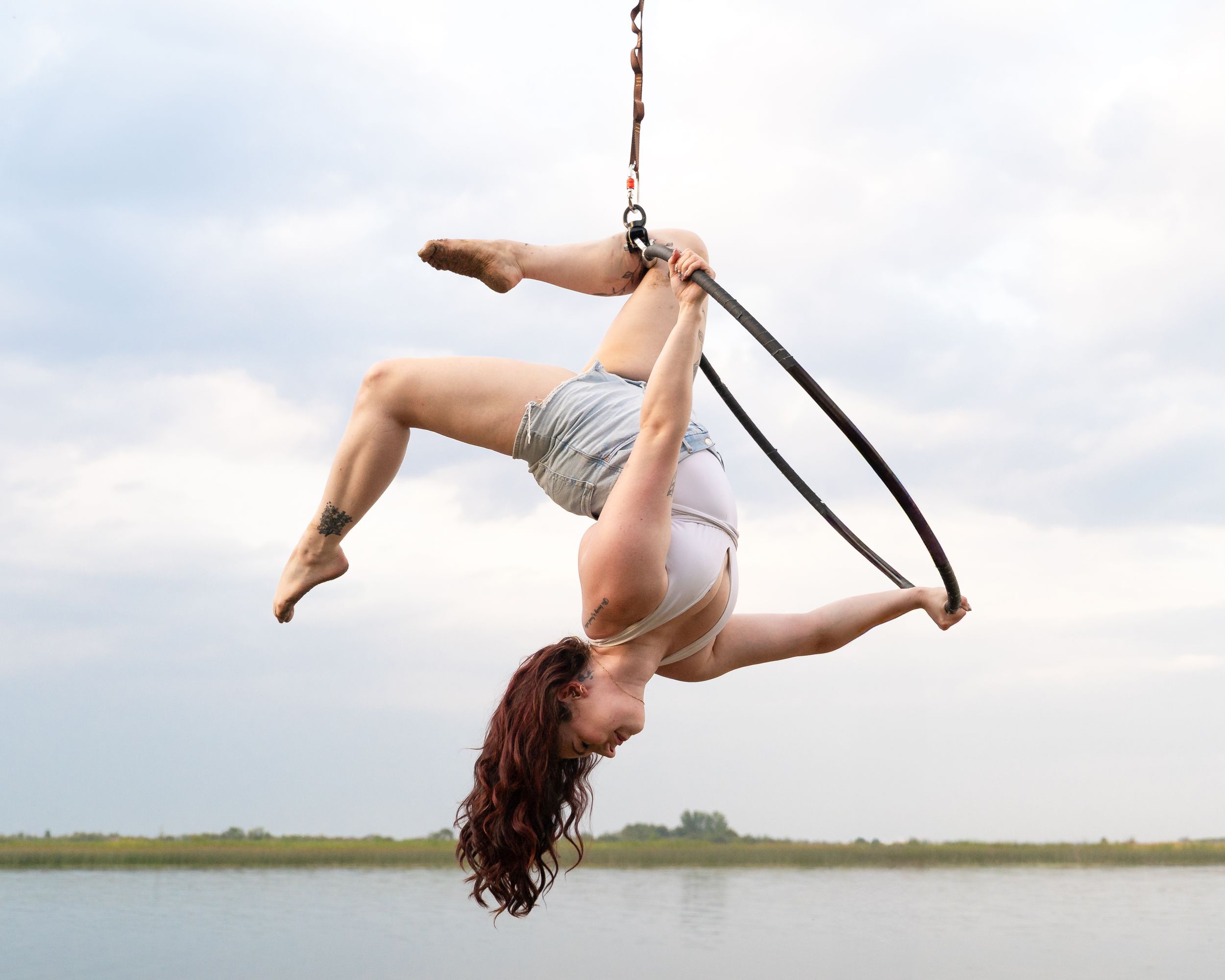 Acrobatic performer executing a dynamic pose while suspended on aerial hoop over water.
