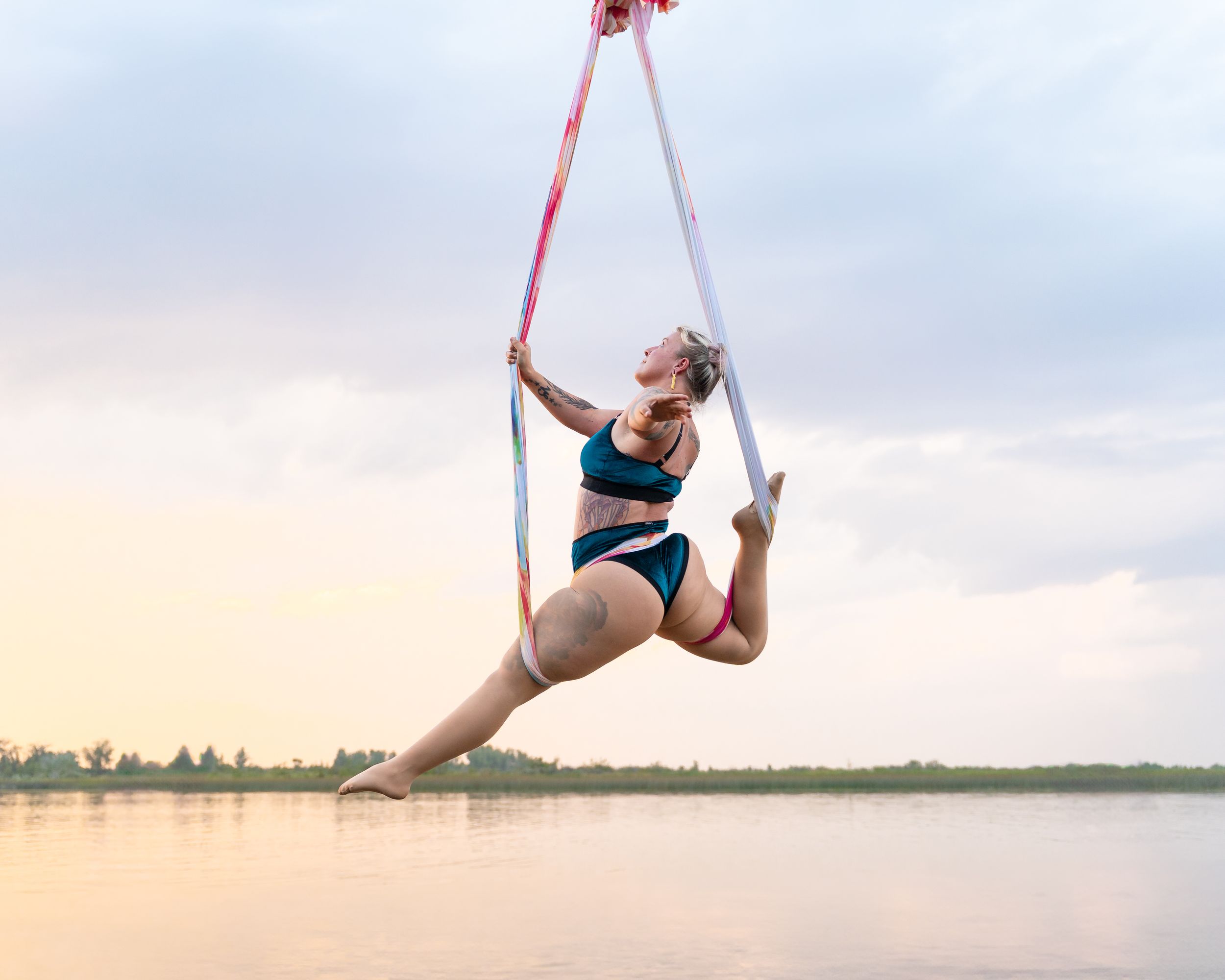 Professional aerial dancer performs graceful splits while suspended from red silk ribbons against cloudy sky.