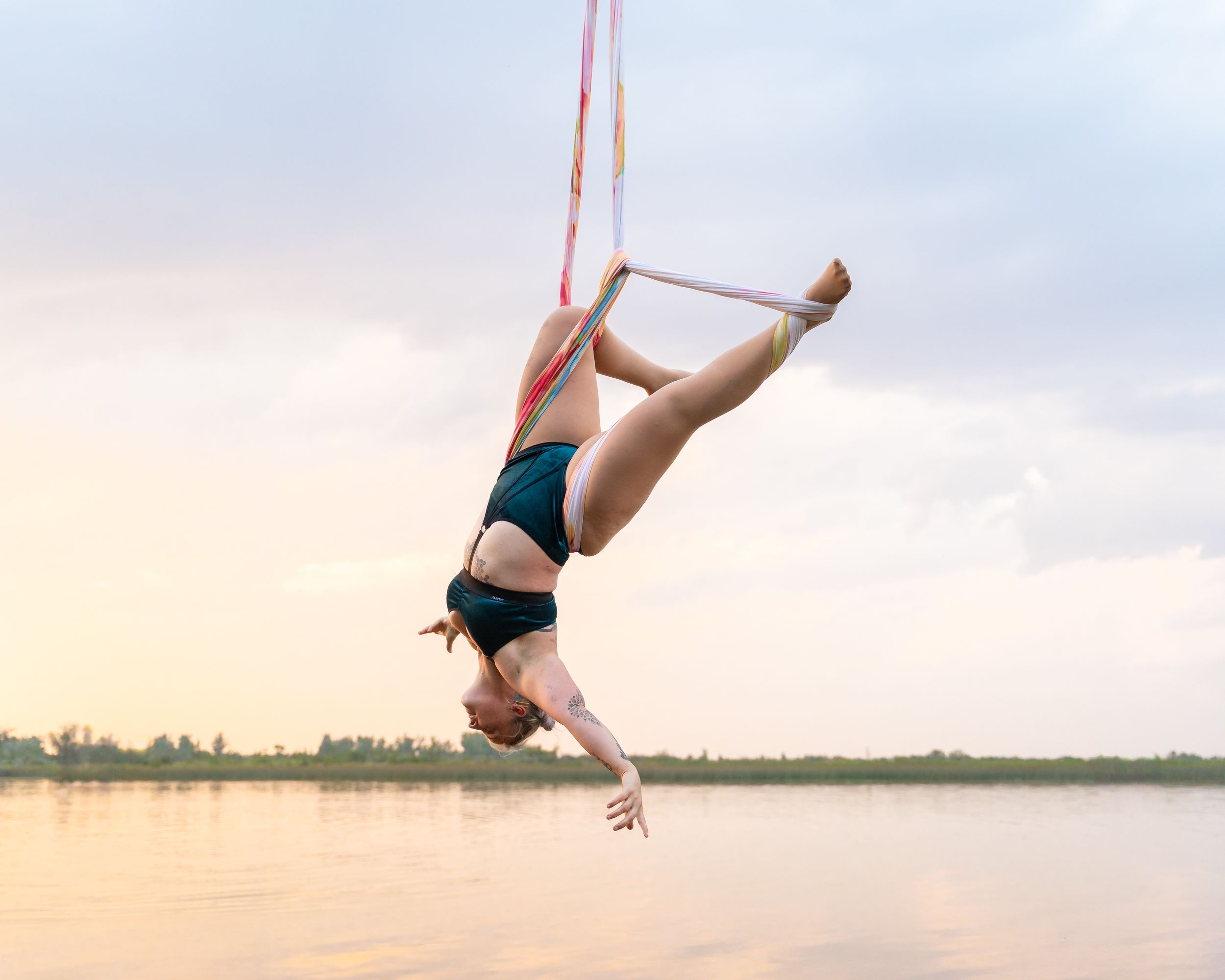 Aerial silks performer gracefully suspended over calm lake waters during sunset in athletic wear.