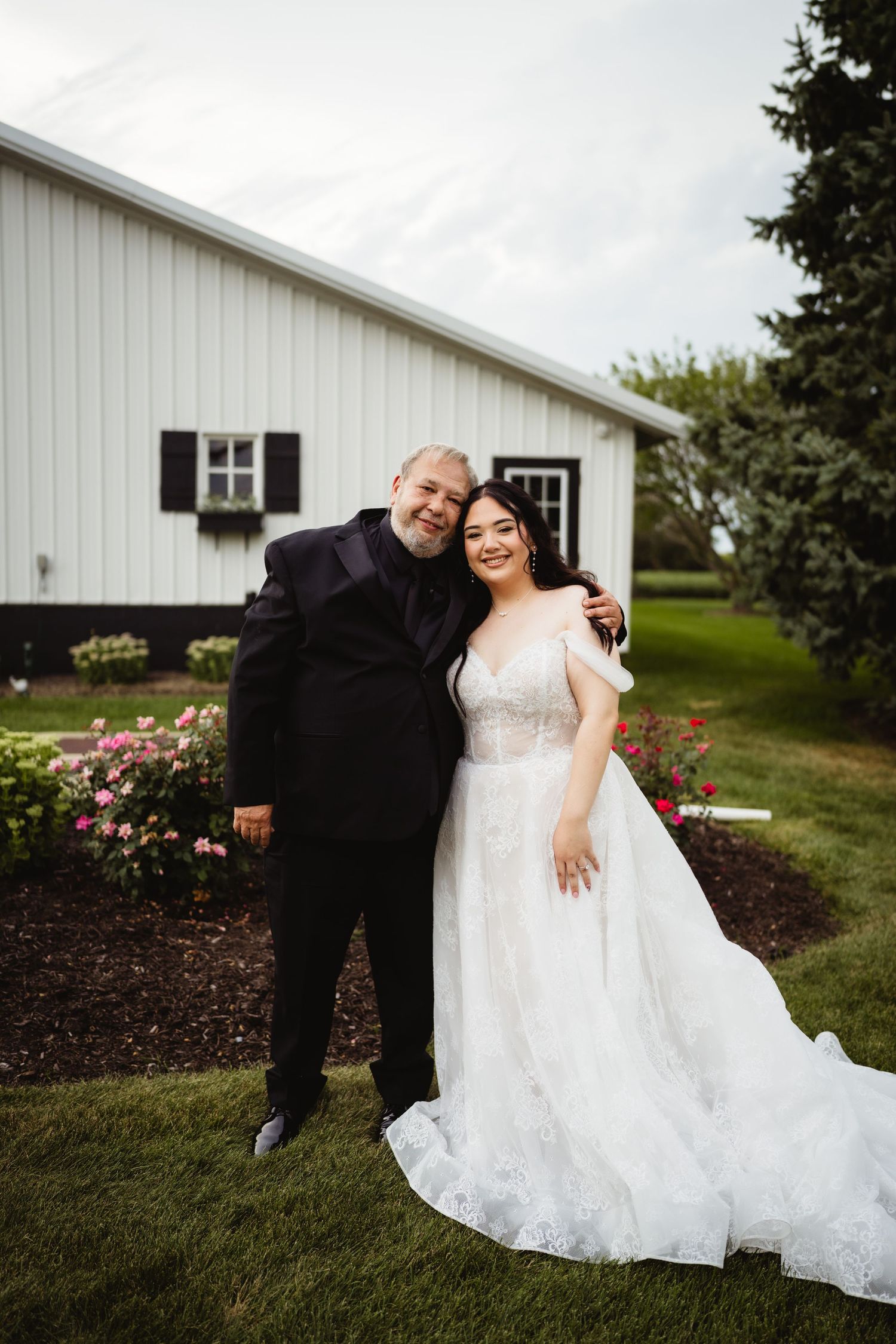 Wedding photo taken outside a white barn with black shutters featuring a couple posing together on the lawn.