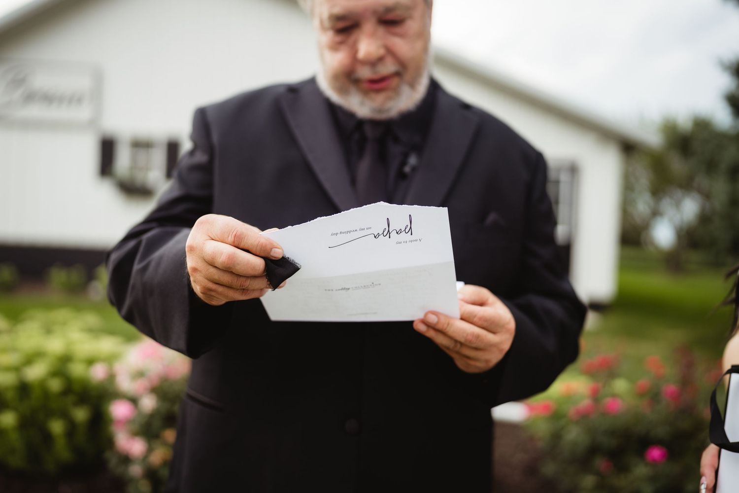 A person in a black jacket reads from an envelope while standing in front of a house with rose bushes.
