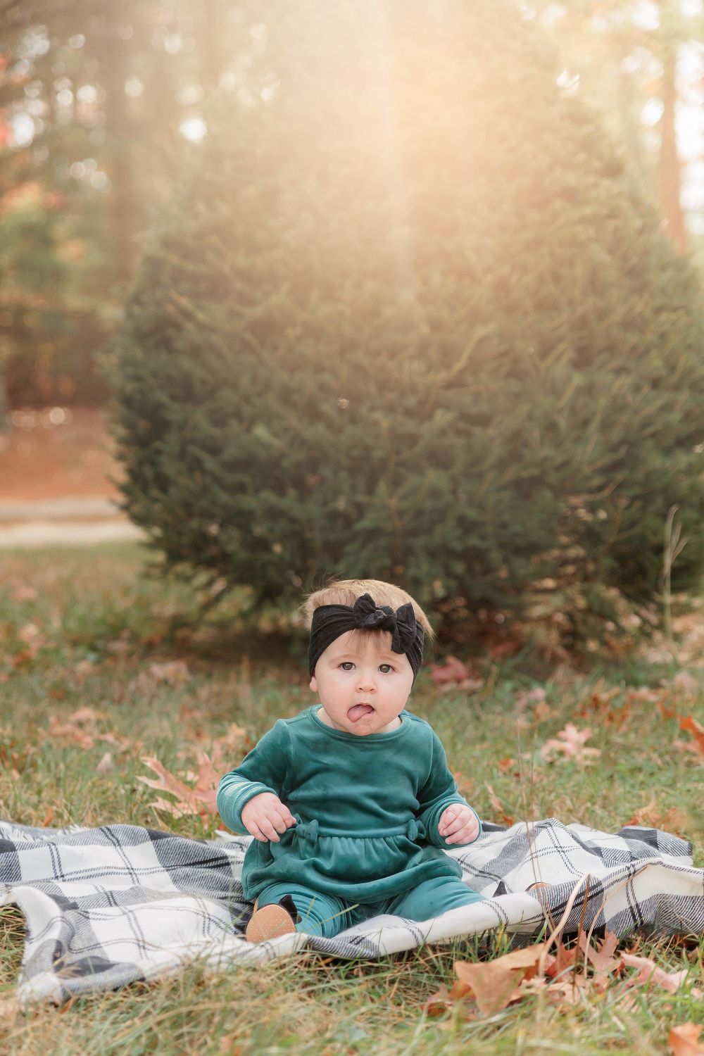 A baby in green sitting on plaid blanket surrounded by evergreen trees and autumn leaves at Turkey Trac Farm in Millville NJ
