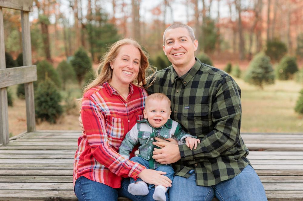 Family wearing red and green plaid shirts sits together laughing on wooden bench during photoshoot at Turkey Trac Farm in Millville NJ
