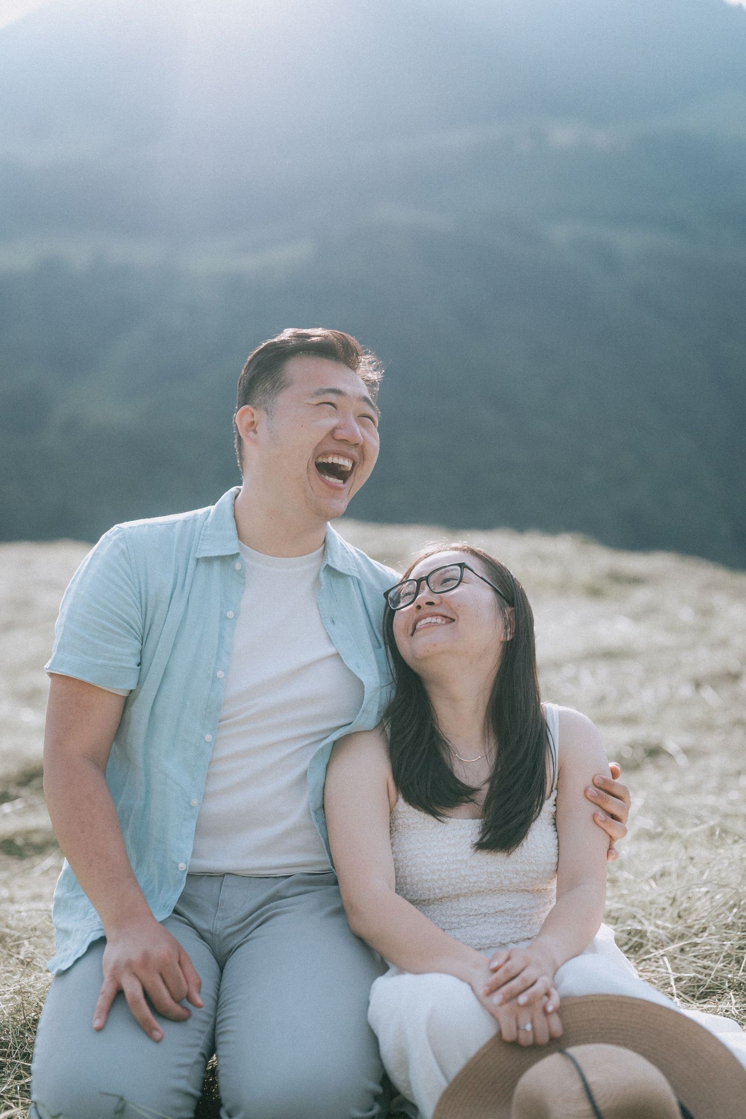 A couple laughs joyfully together while sitting outdoors on a misty mountaintop with scenic views in the background.