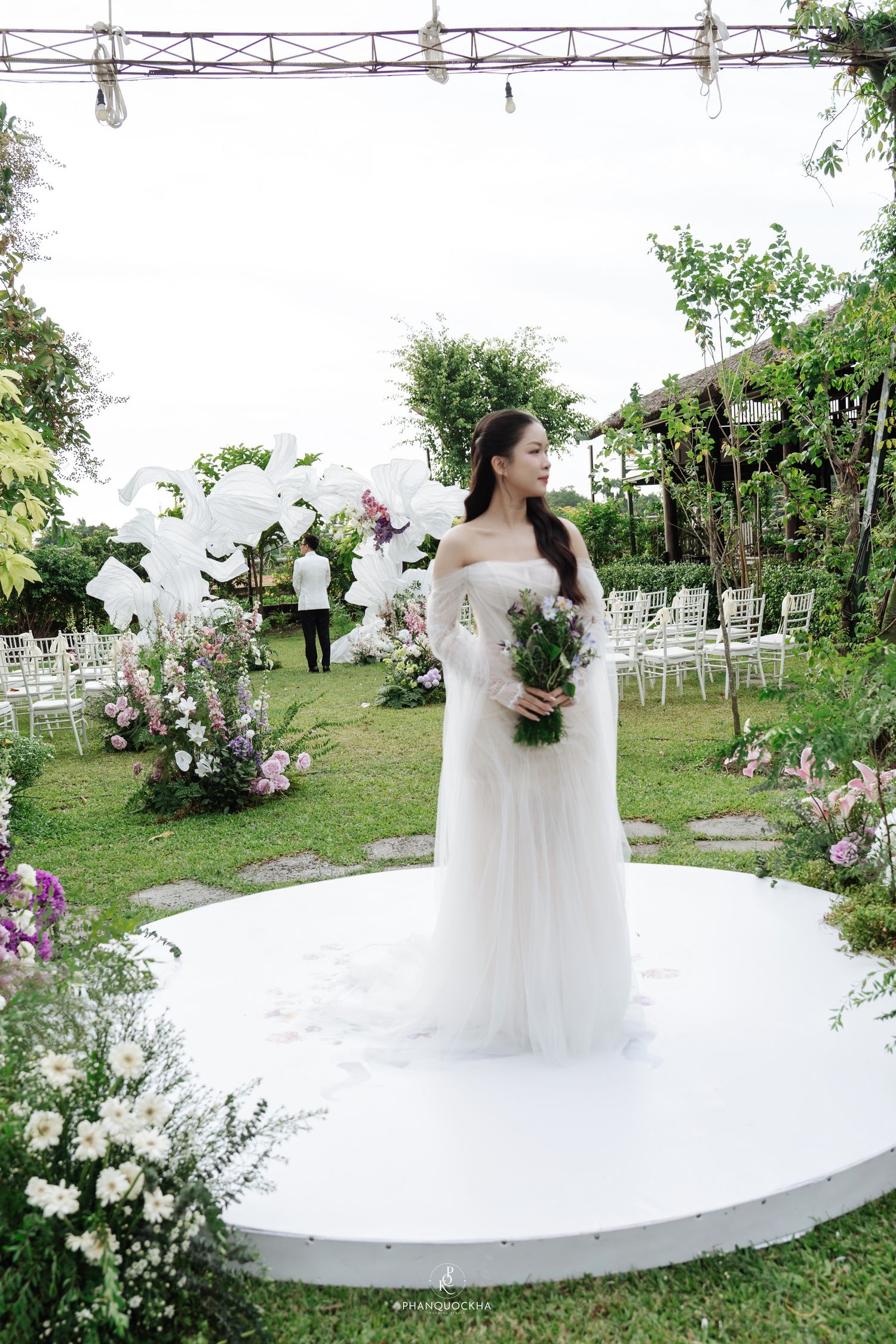Bride in white off-shoulder gown standing on circular platform at outdoor garden wedding.