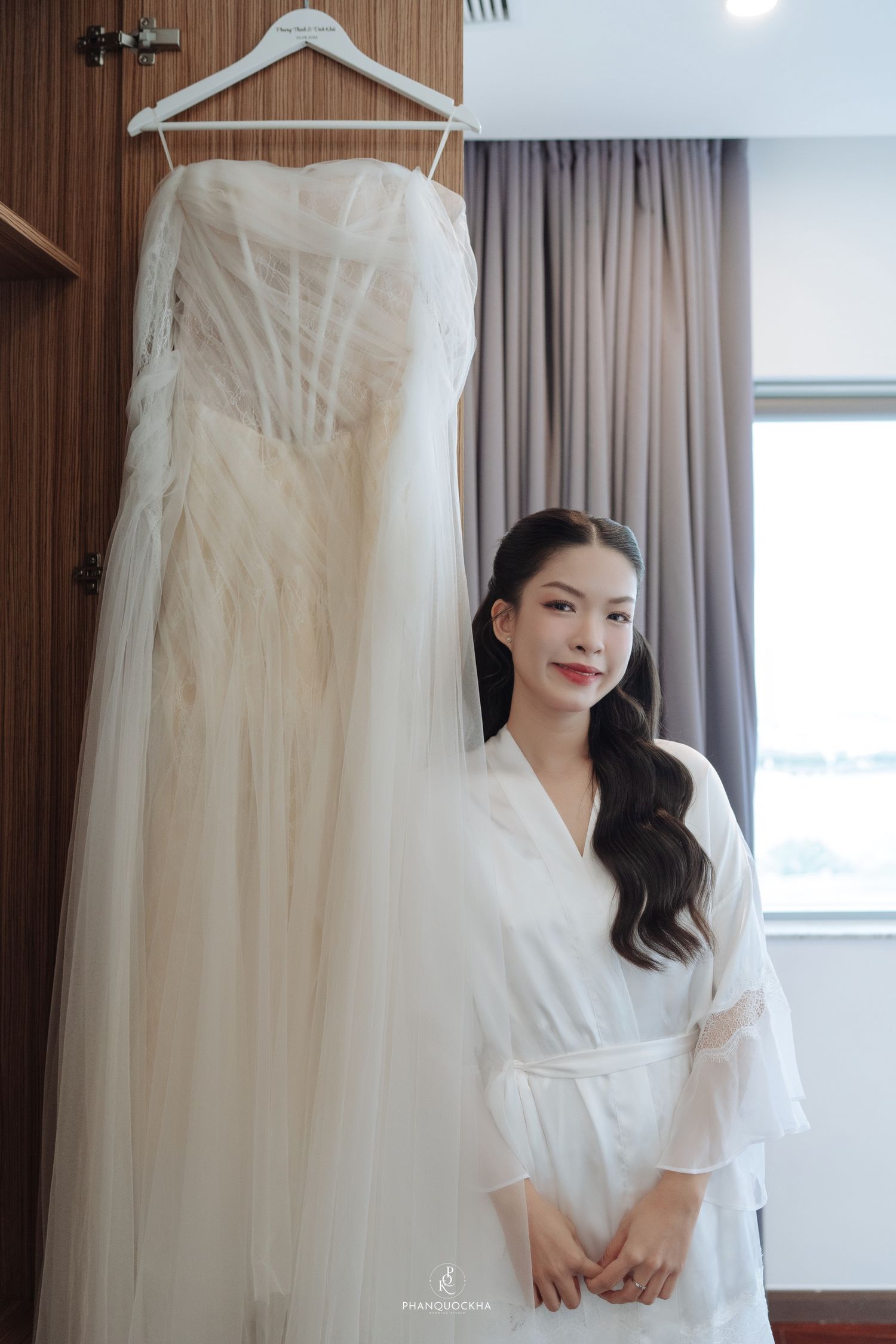 Smiling Asian bride in white robe standing beside her elegant strapless tulle wedding dress.
