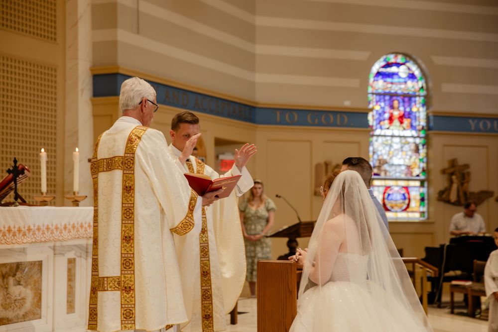 A wedding ceremony takes place at a church altar with religious officiants and a stained glass window in the background.