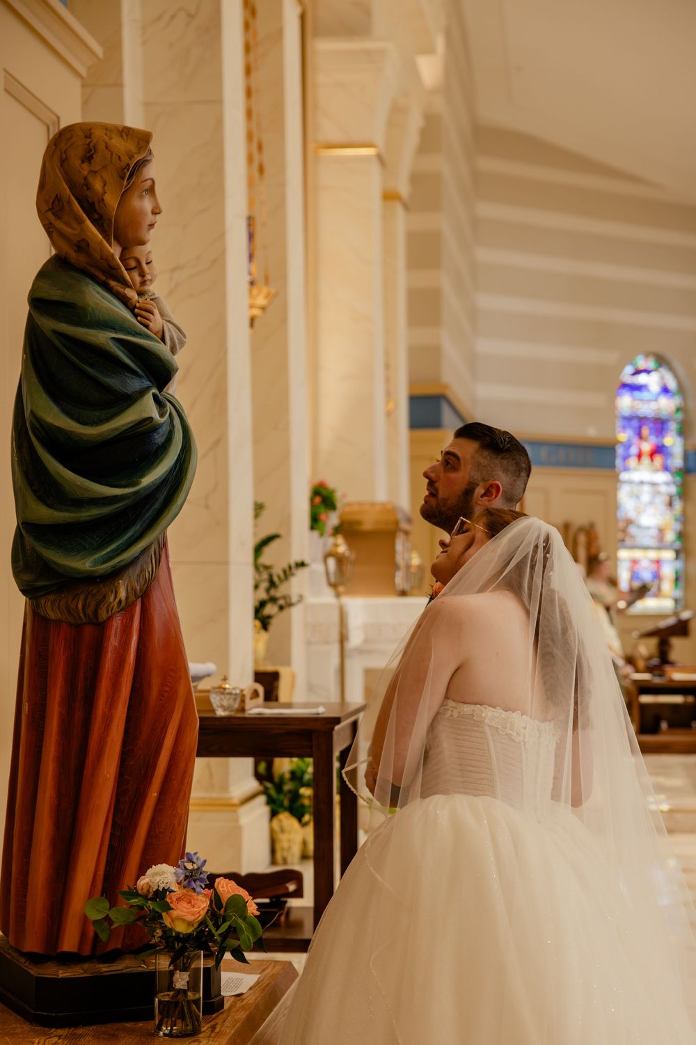 A person in a wedding dress kneels before a religious statue in a church with stained glass windows in the background.