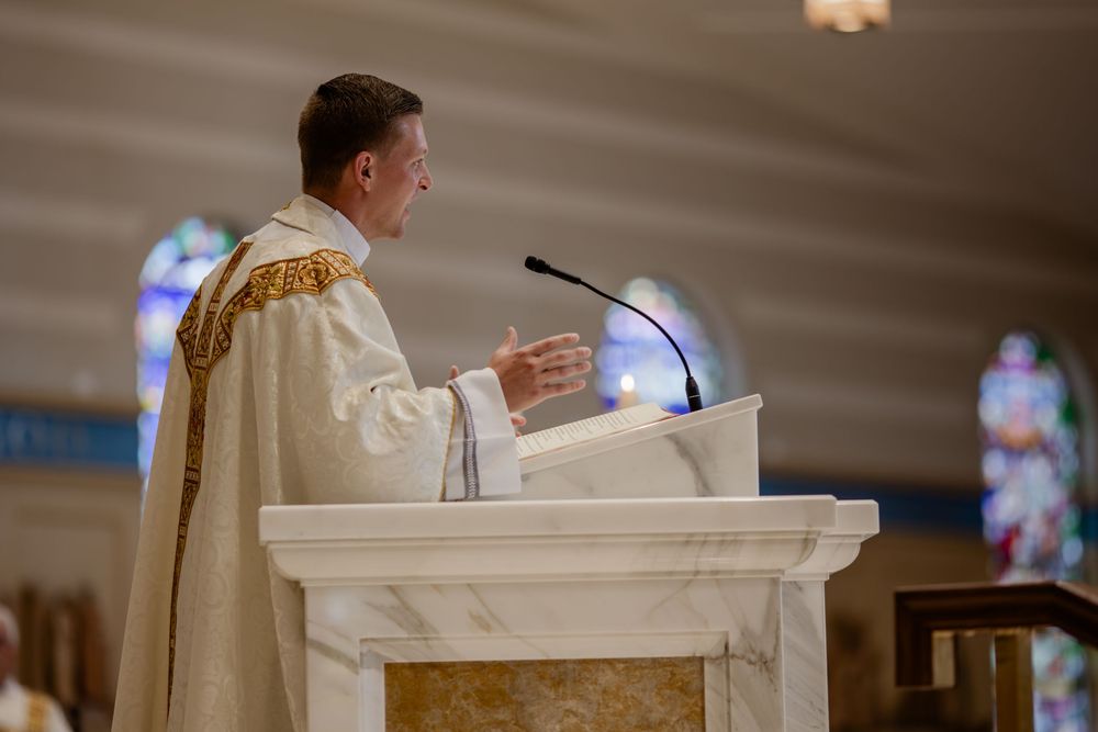 A clergy member in cream colored vestments delivers a sermon from a white marble pulpit in a church.