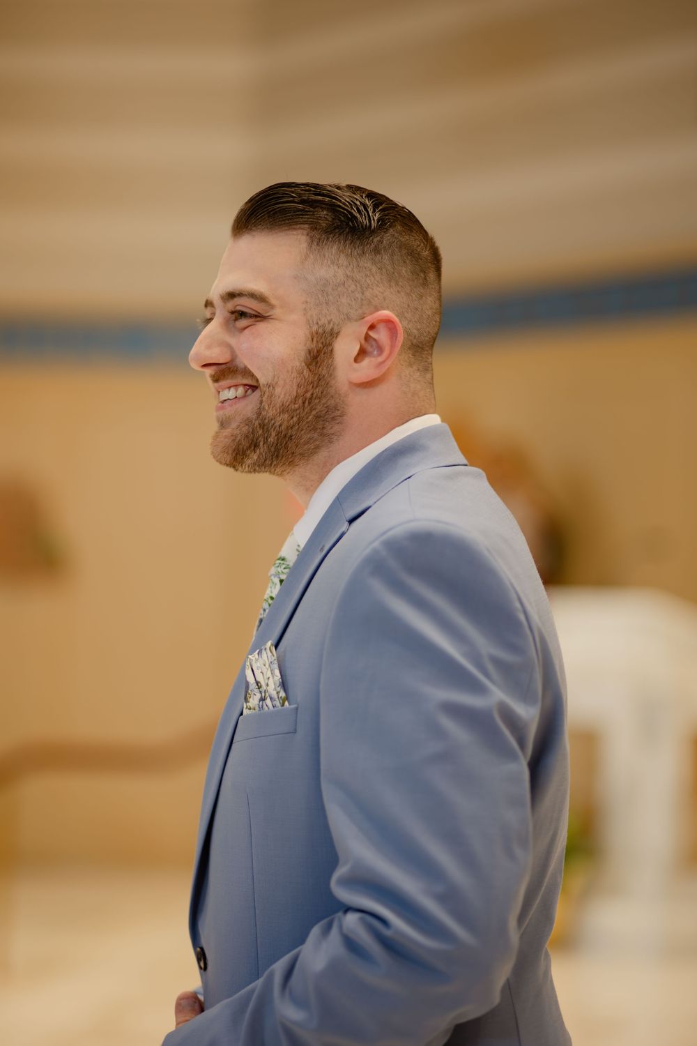 A happy groom in a light blue suit smiles warmly during a wedding celebration.