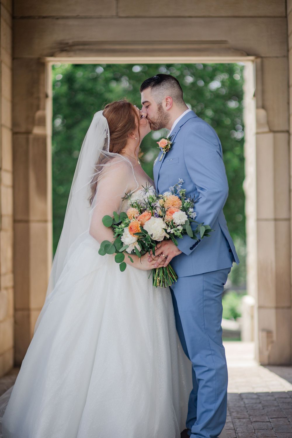A romantic wedding kiss between newlyweds under an archway, with the bride holding a peach and white floral bouquet.