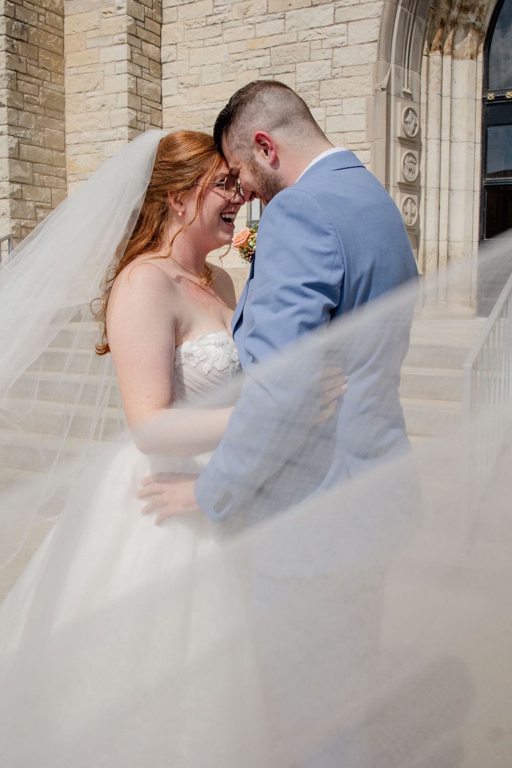 Romantic moment between newlyweds against stone church wall with flowing veil in sunlight.