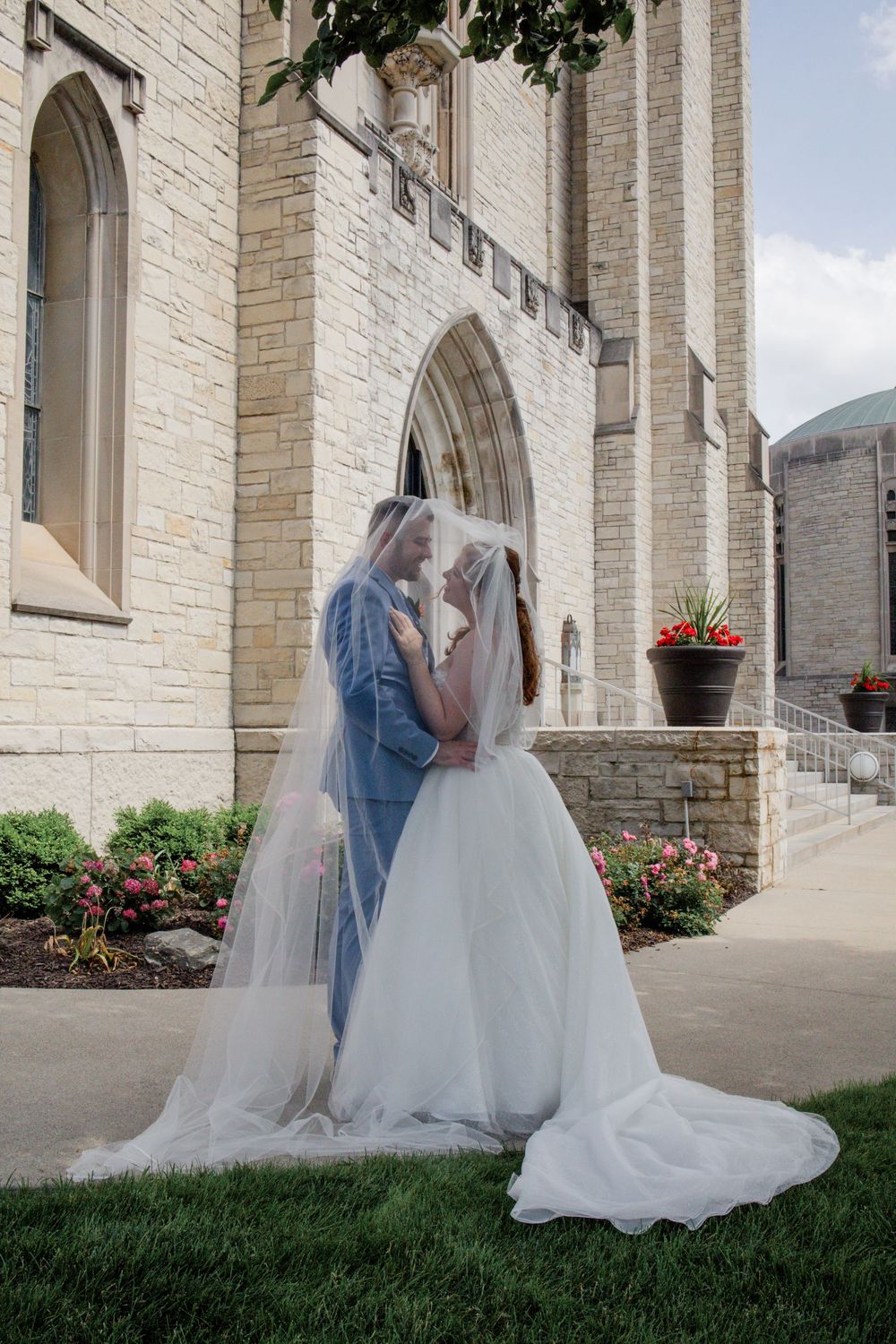 Wedding couple embracing outside stone church with arched doorways and flowers.