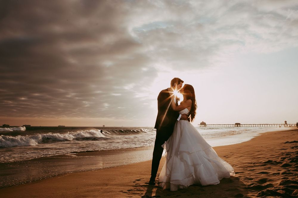 A couple stands on a dreamy beach at sunset with dramatic clouds above in a panoramic wedding photograph in Laguna Beach.