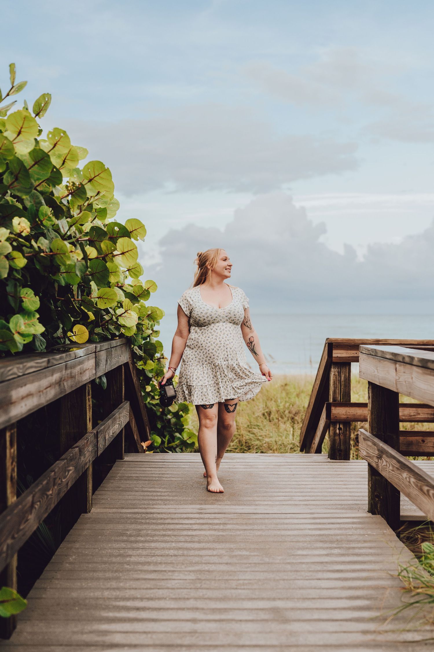 Figure in a polka dot dress walks along a wooden boardwalk overlooking beach scenery on a cloudy day.