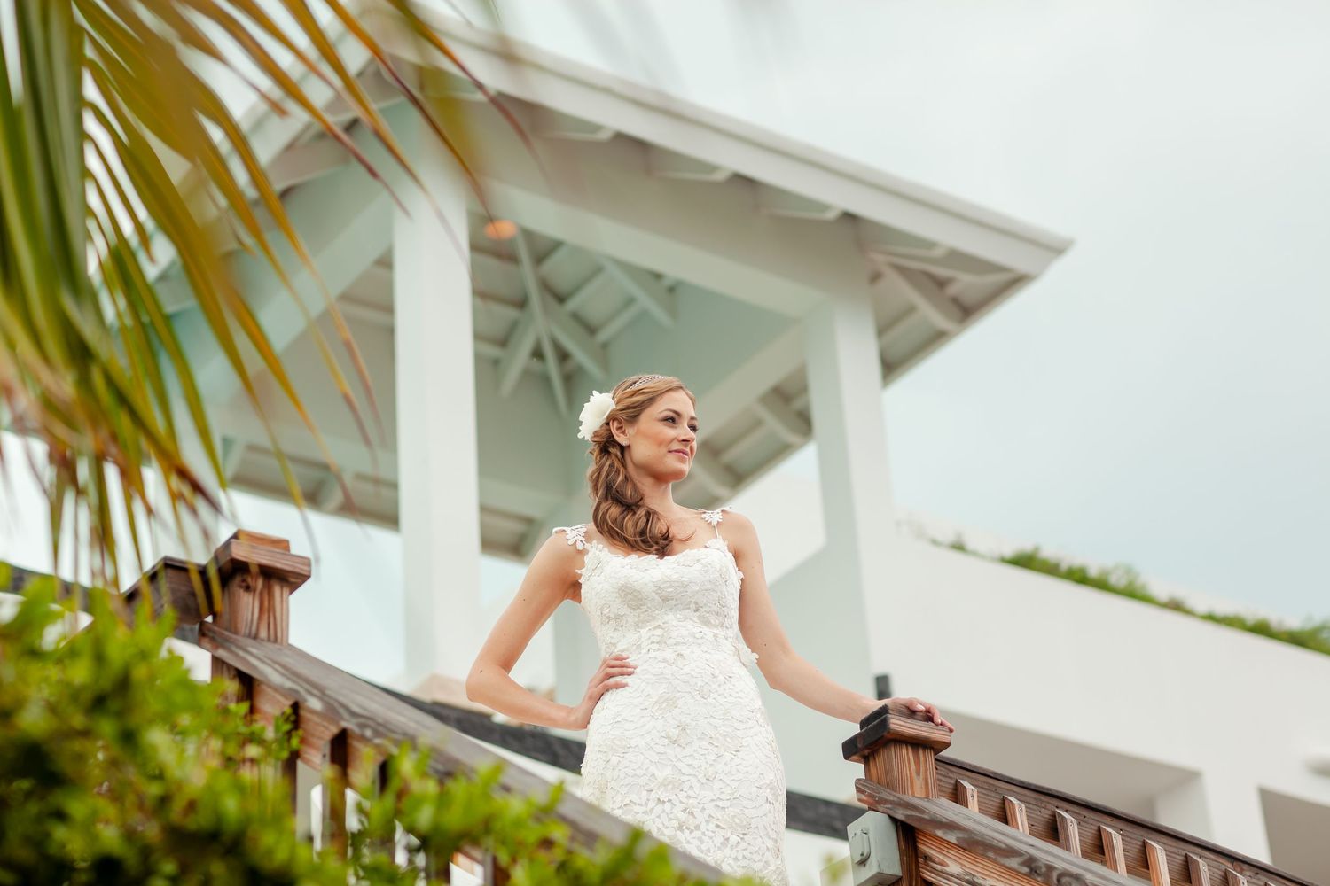 Bride in white gown stands on balcony overlooking tropical landscape with palm trees.