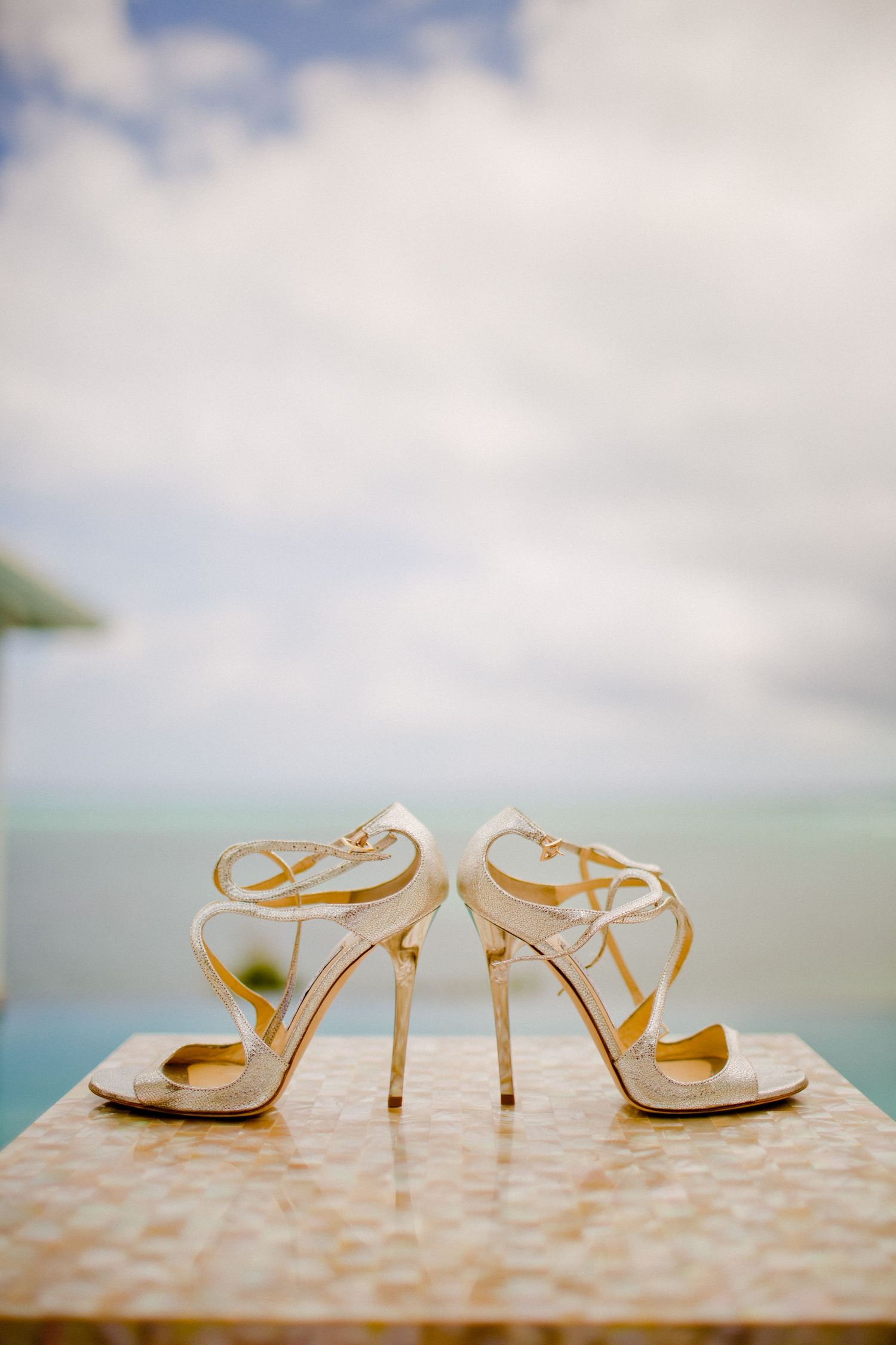 Elegant white strappy bridal sandals with high heels photographed against ocean backdrop.