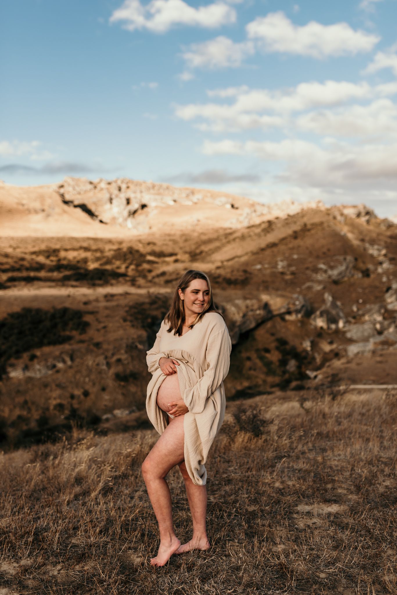 Expecting mother in flowing dress stands barefoot on hillside during golden hour in Christchurch.