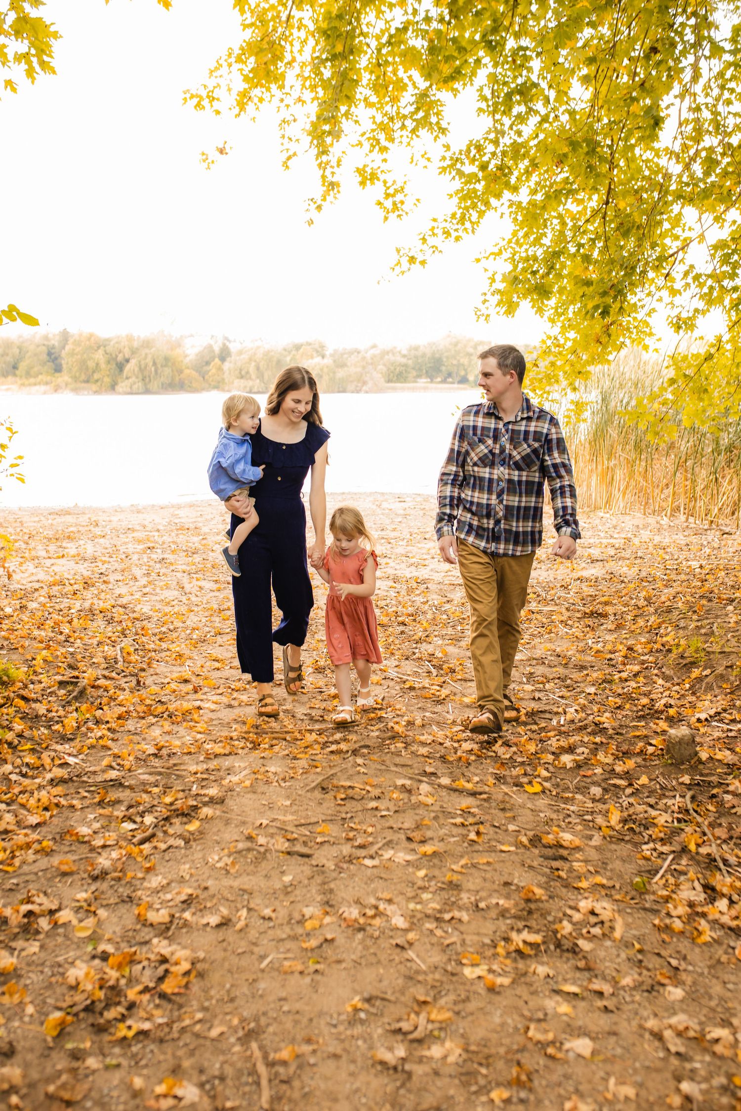 A family walks together on a leaf-covered path by a lake during autumn with golden trees overhead.