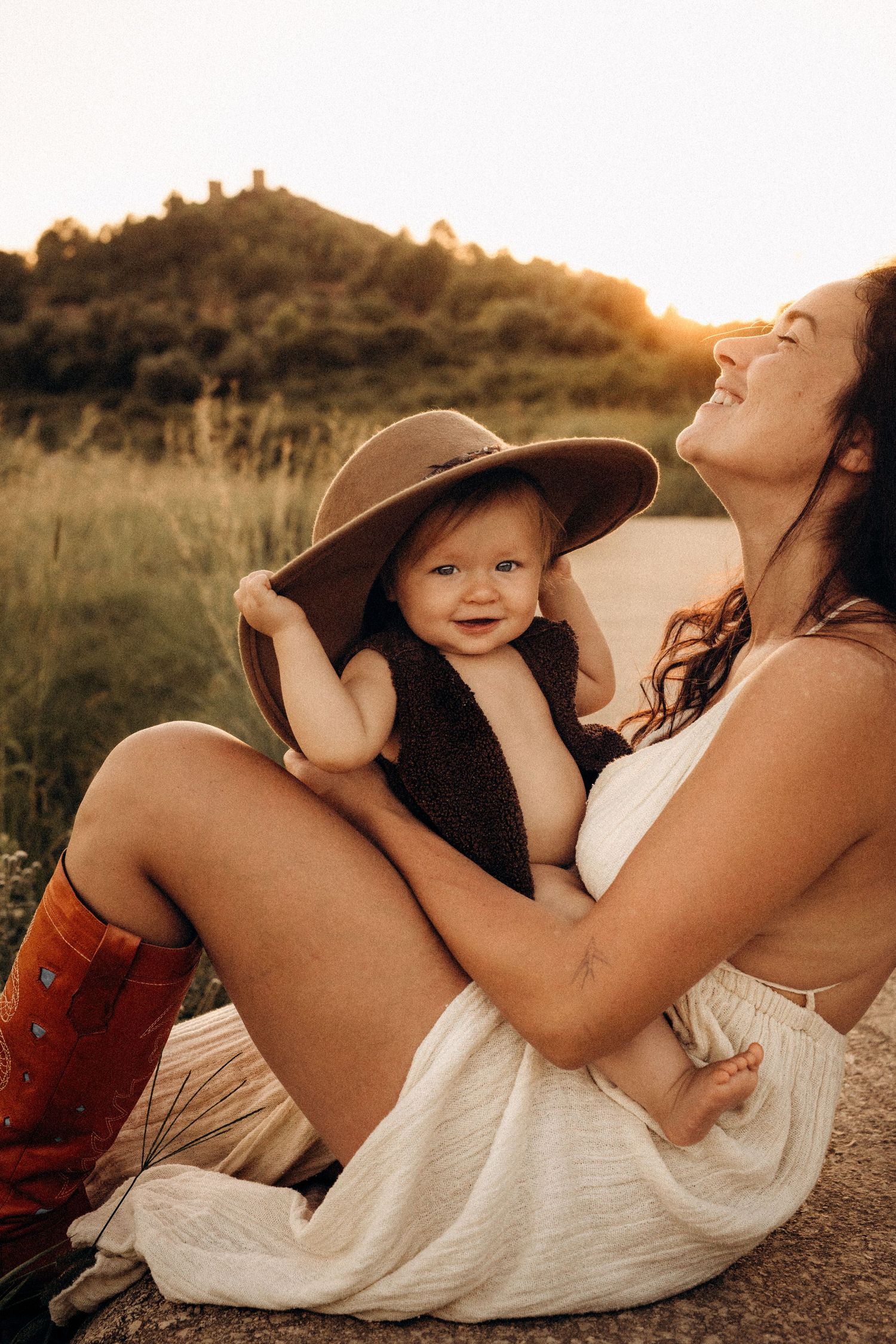 A parent and baby in a brown hat share a tender moment outdoors during golden hour sunset. Javea photographer