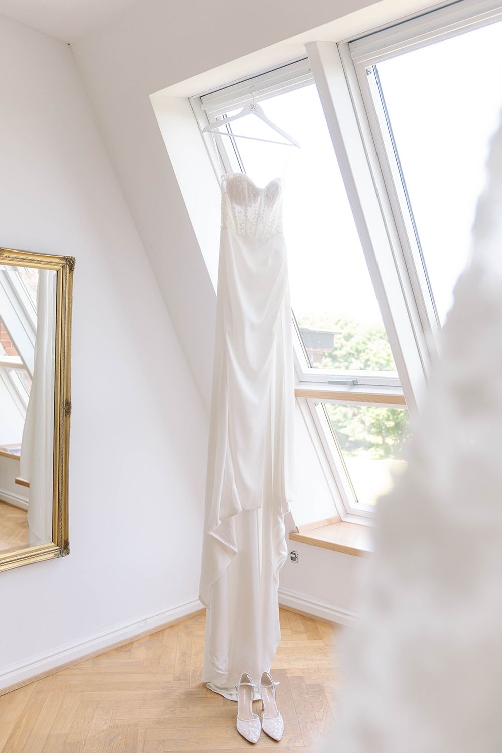 White wedding dress hanging elegantly by a skylight window in a bright room.