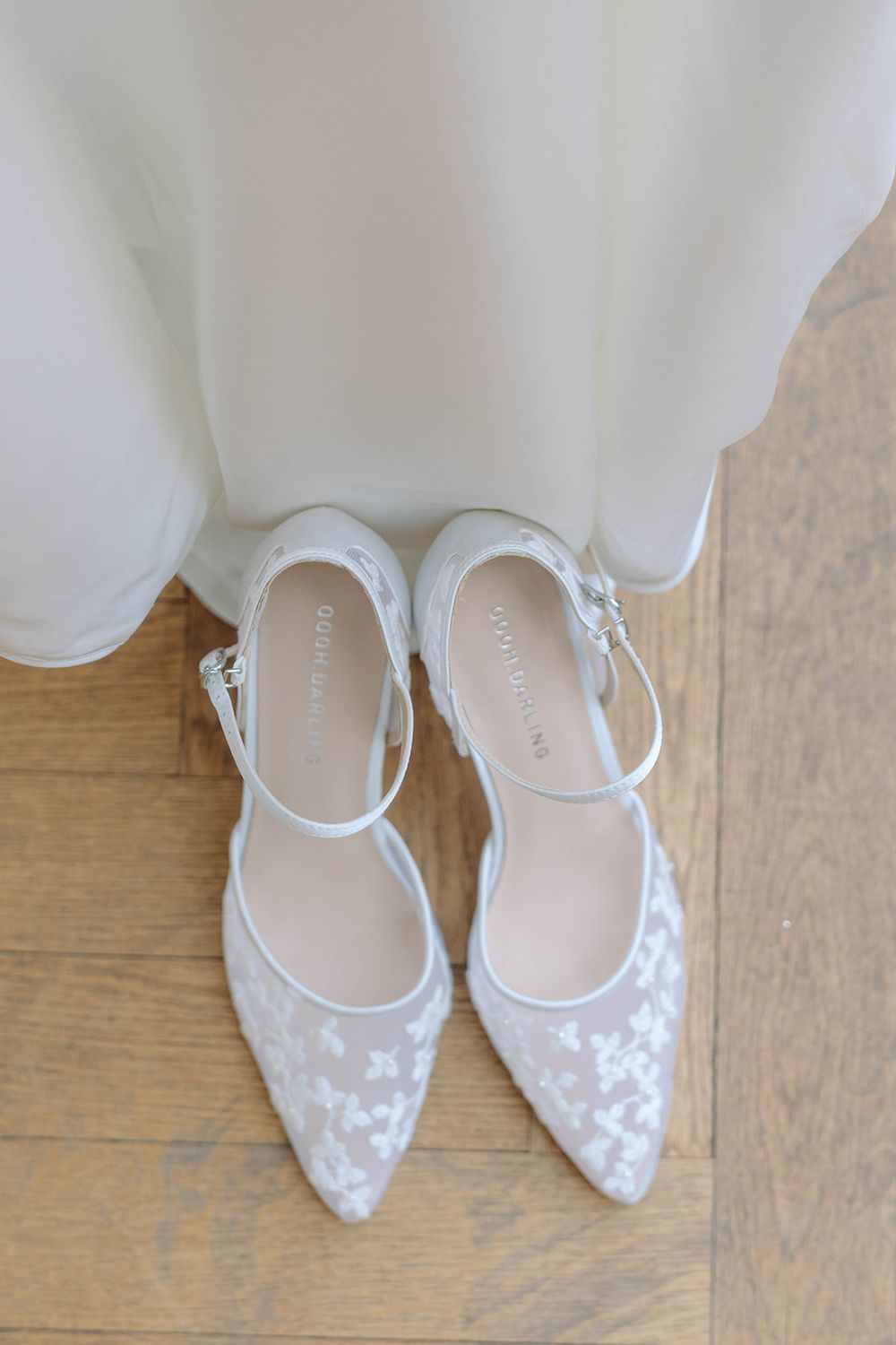 Delicate white lace bridal shoes with ankle straps displayed against a wooden floor.