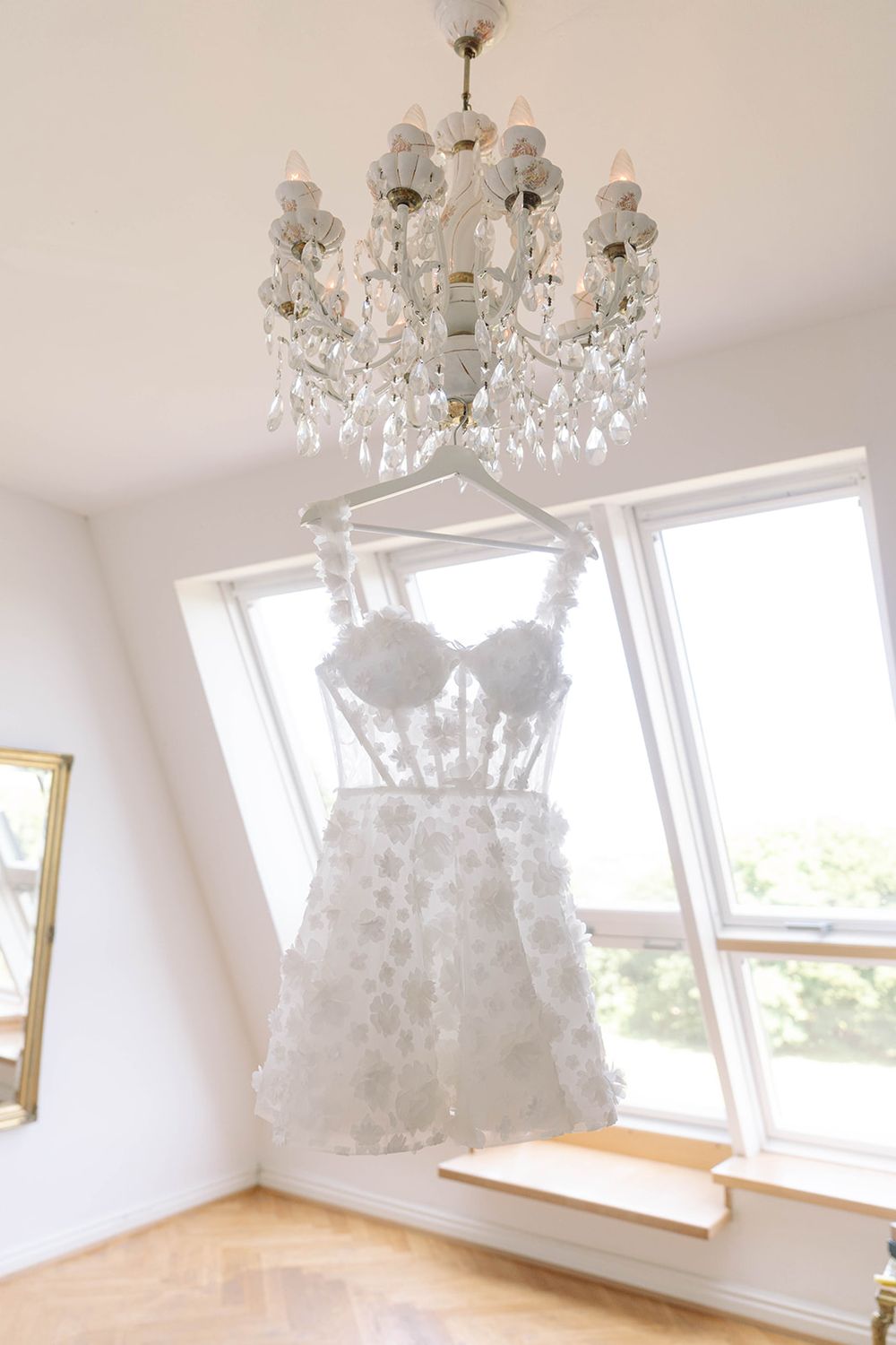 Elegant white wedding dress hanging beneath a crystal chandelier in a sunlit room.