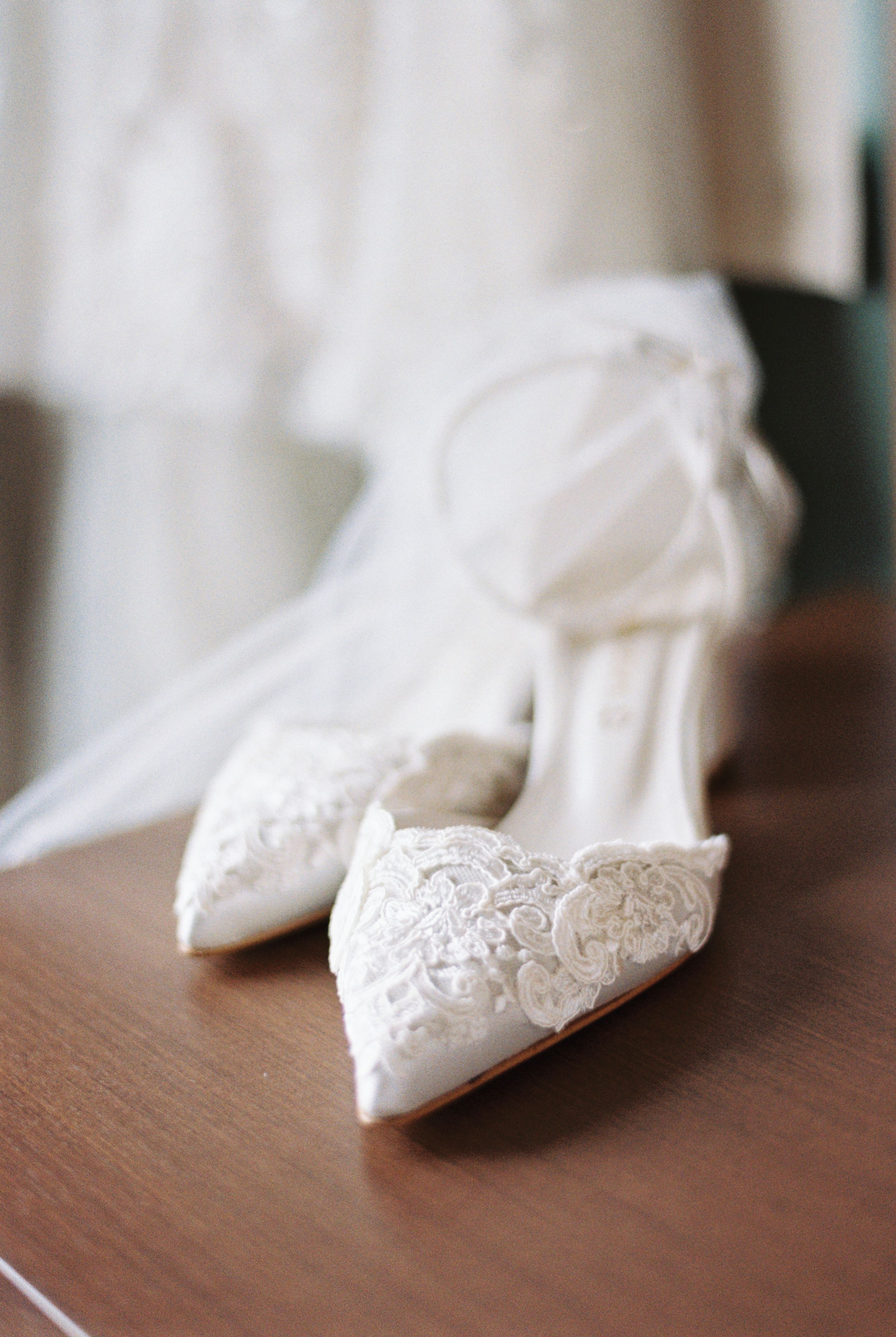 Elegant white lace wedding shoes with pointed toes and delicate detailing sit on a wooden surface.