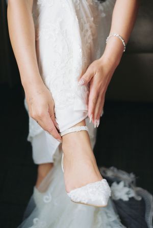 Close-up of hands helping a bride put on delicate white lace wedding shoes with a flowing dress in the background.