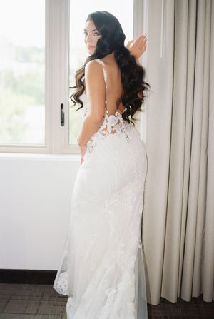 Back view of an elegant white lace wedding dress against a window and curtained backdrop.
