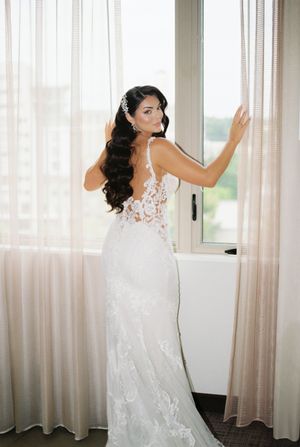 Bride in lace wedding dress poses playfully near curtained windows in a bright room.