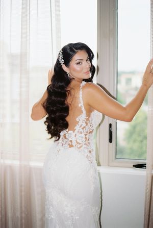 A bride in an elegant lace wedding gown with flowing dark curls stands gracefully by a window.