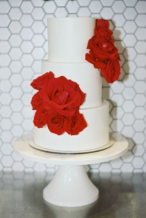White tiered wedding cake decorated with vibrant red roses against a modern hexagonal tile background.