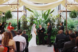 Indoor wedding ceremony taking place in a bright greenhouse conservatory filled with lush tropical plants and white drapery.