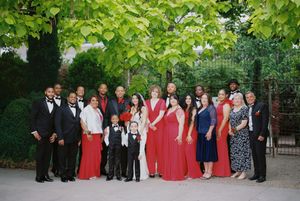 Wedding party poses for a formal group photo in red and black attire under green trees.