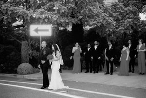 Black and white wedding processional photo showing bridal party walking down outdoor path with guests watching.