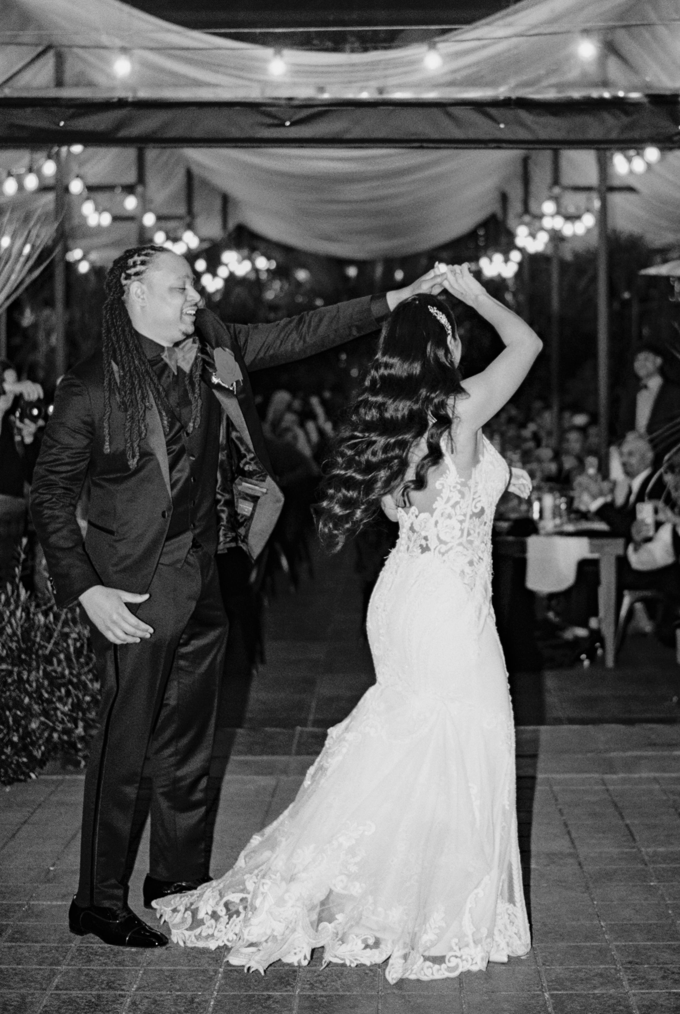 Black and white photo of newlyweds dancing together under outdoor tent lighting.