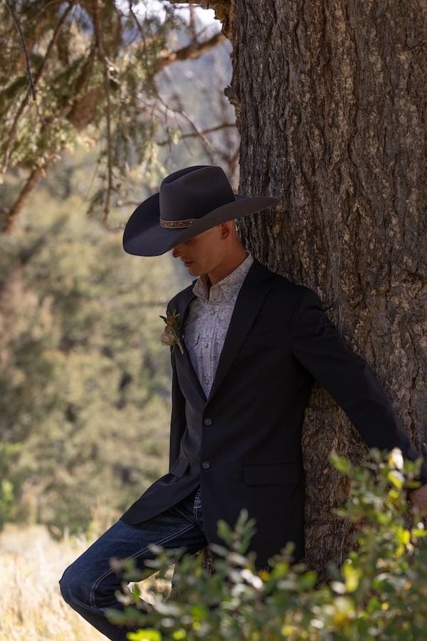 Black-clad figure leans against a large tree trunk in a natural outdoor setting with filtered sunlight.