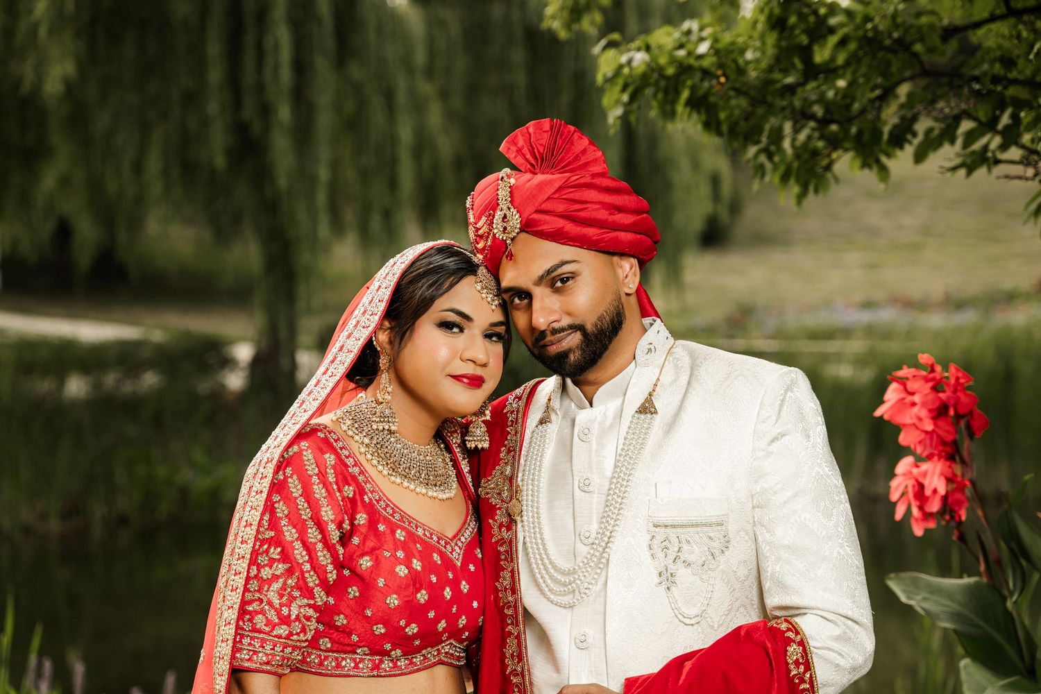 Wedding couple in red and white traditional Indian attire pose by lake with willow tree.