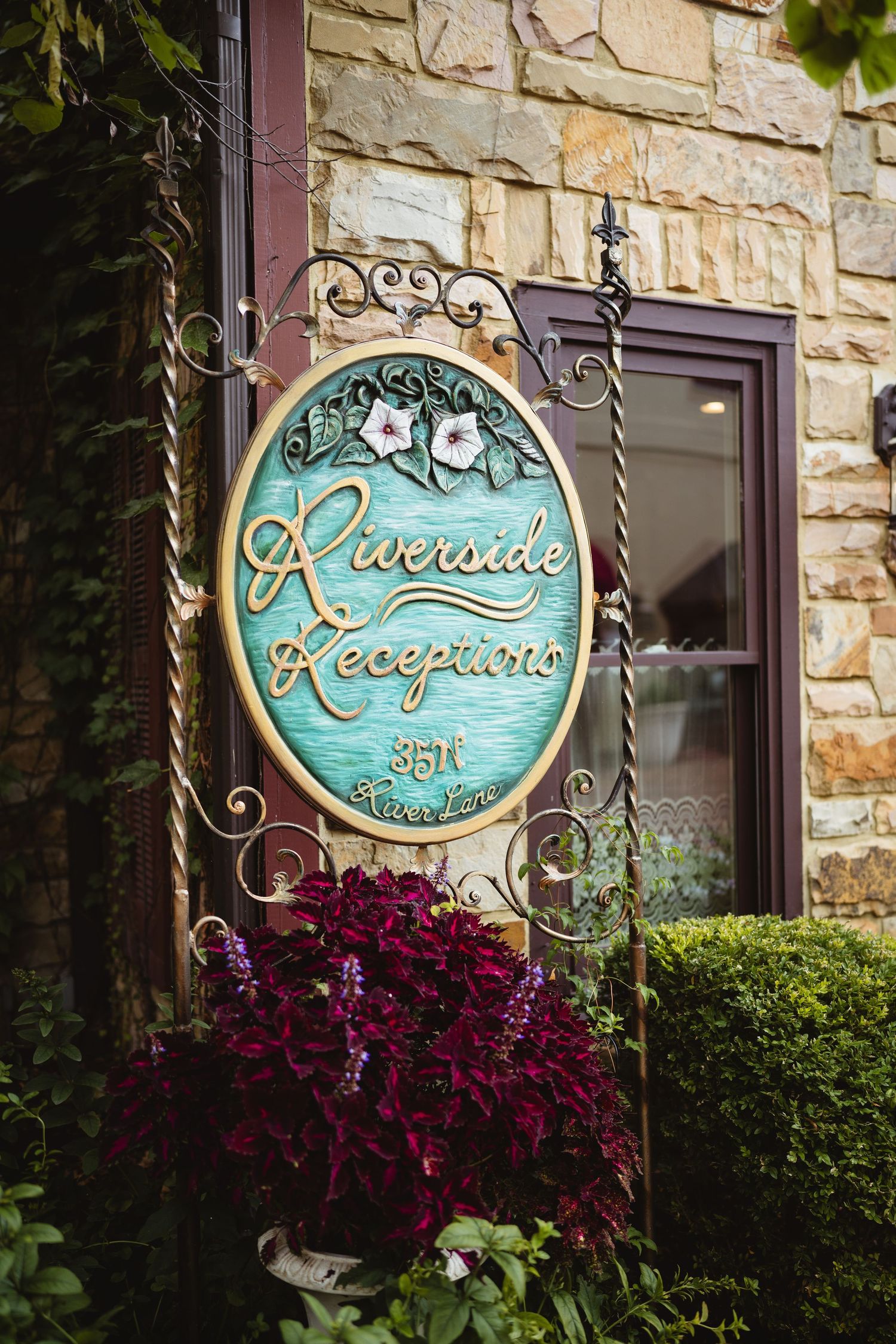Elegant turquoise circular business sign with decorative metalwork and red flowers blooming below against a stone wall exterior.